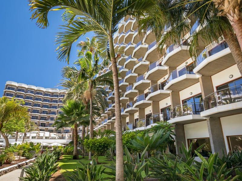 Modern hotel complex with balconies and palm trees under a clear blue sky in daylight.
