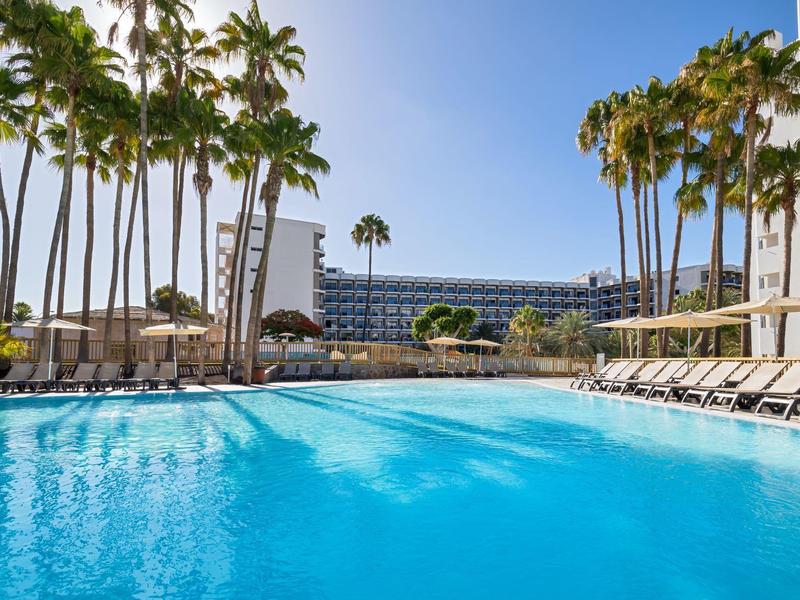 Large pool area with palm trees, lounge chairs, and a hotel building in the background.