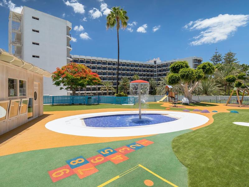 Hotel with swimming pool, water feature, and play area surrounded by palm trees under blue sky.