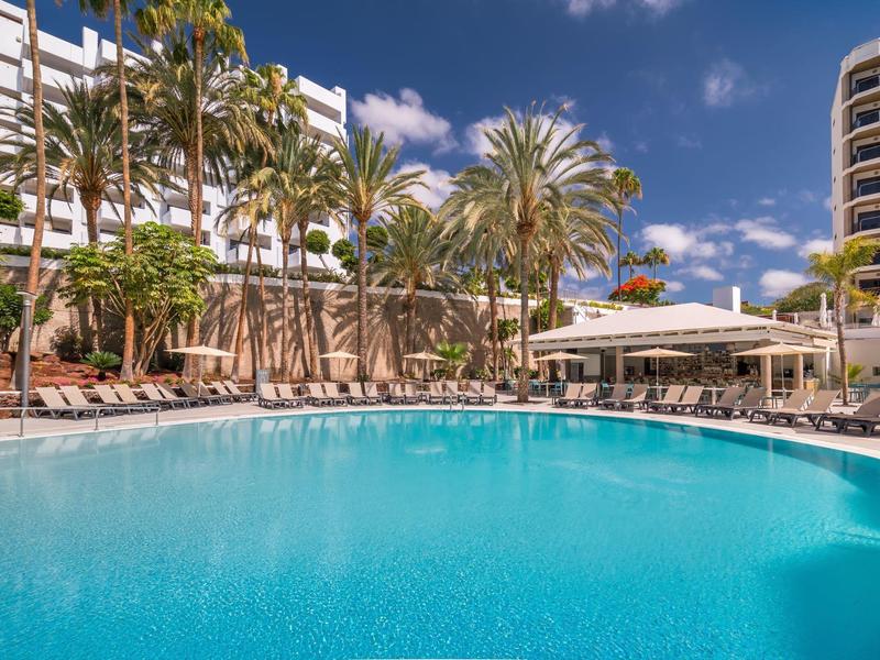 Large outdoor pool with lounge chairs and palm trees in front of hotel buildings under a sunny sky.