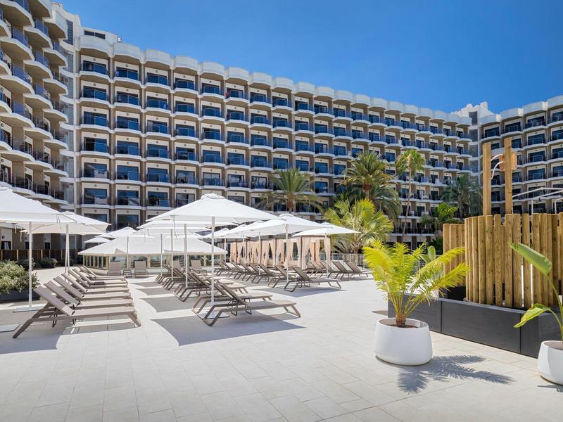 Hotel with outdoor pool, sun loungers, and umbrellas under blue sky.