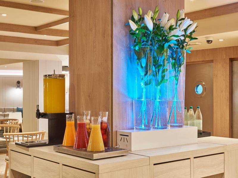 Hall di un hotel moderno con vasi illuminati, fiori freschi e succhi di frutta su un mobile in legno.