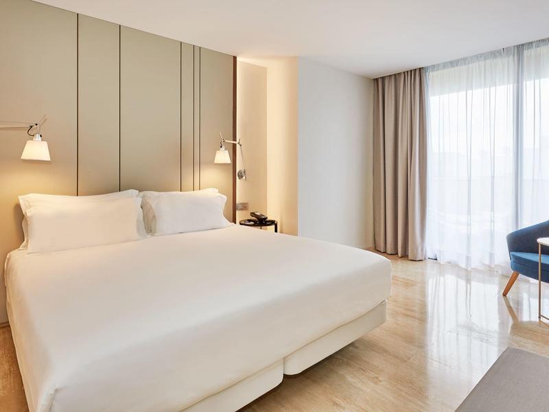 Camera d'hotel luminosa con letto grande, comodini e poltrone vicino a finestre a tutta altezza.