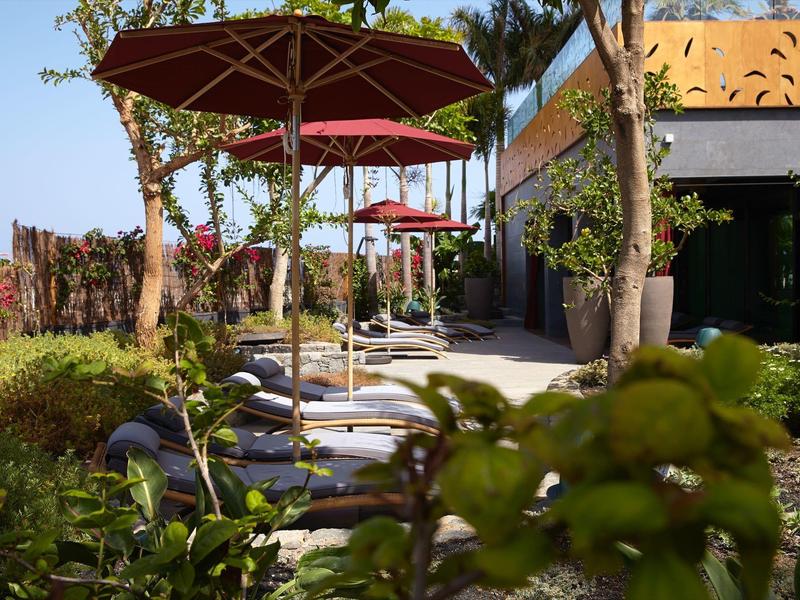 Giardino con lettini e ombrelloni rossi accanto a un edificio moderno con tempo soleggiato.
