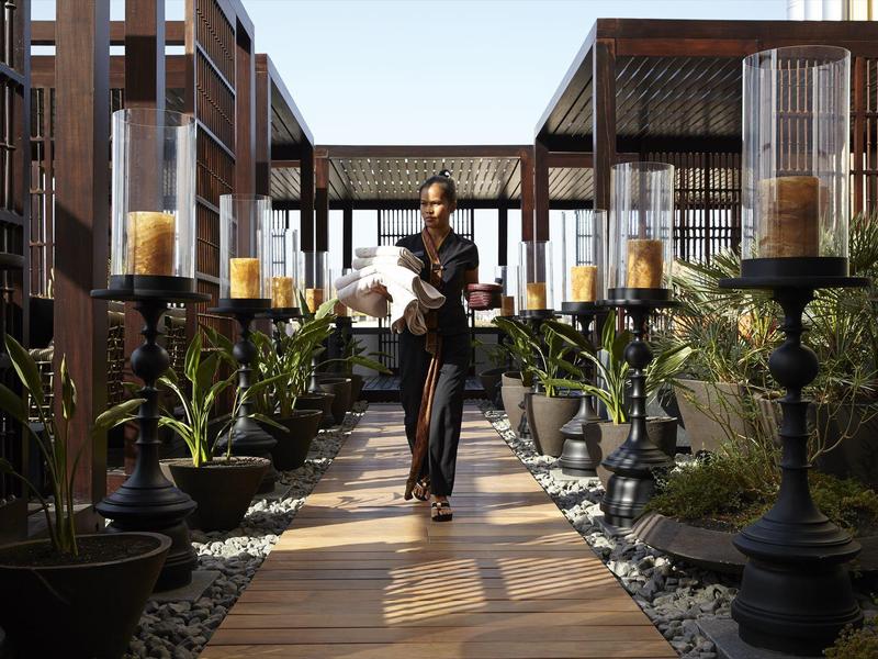 Personale dell'hotel con vassoio che cammina su un sentiero in legno tra piante e lanterne.