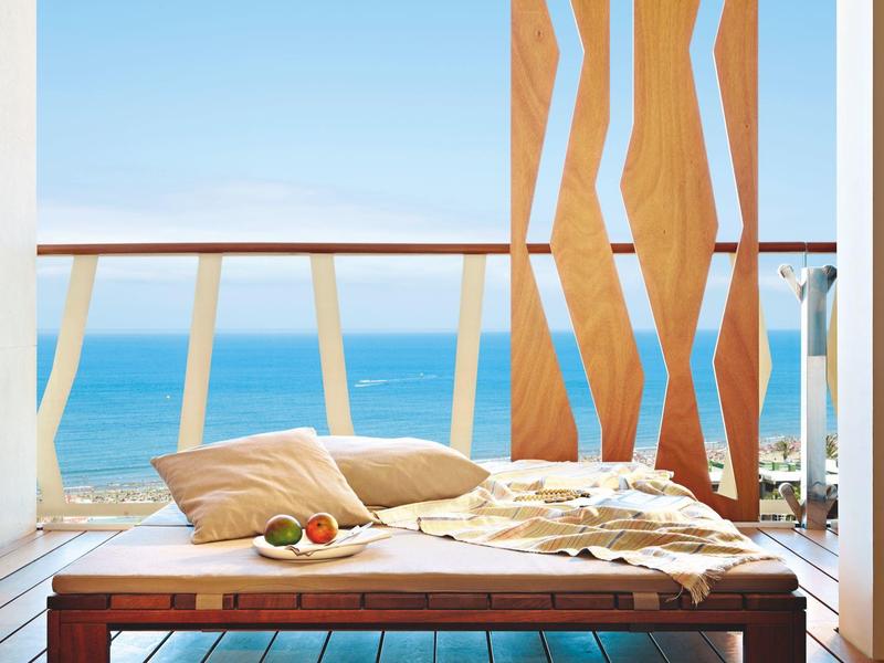 Terrazza con sdraio in legno, cuscini e vista sul mare blu sotto un cielo sereno.