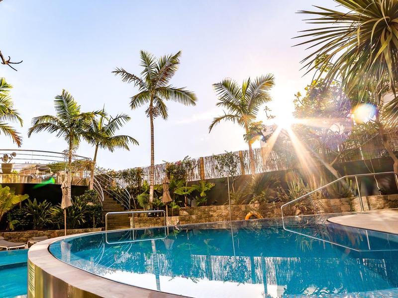 Area piscina dell'hotel con palme e vista sulla città al tramonto.
