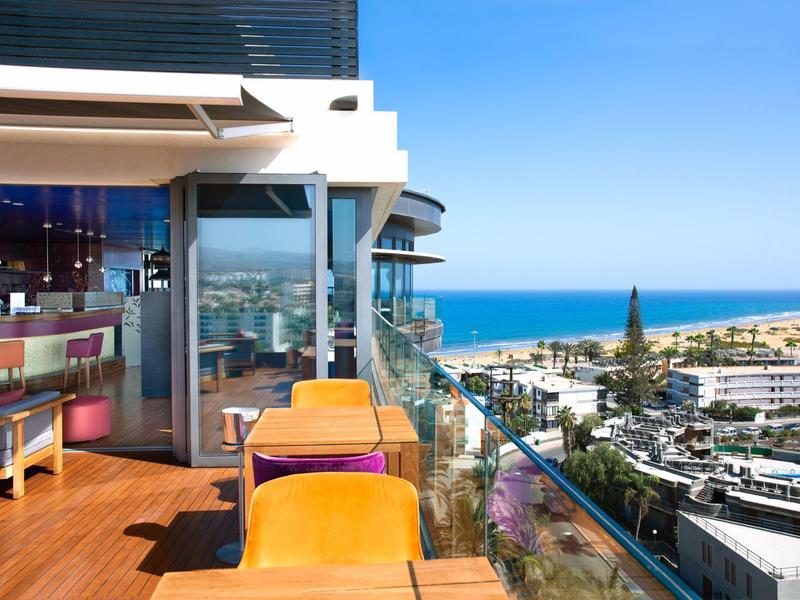 Balcone con tavoli e sedie con vista sul mare e sugli edifici circostanti.