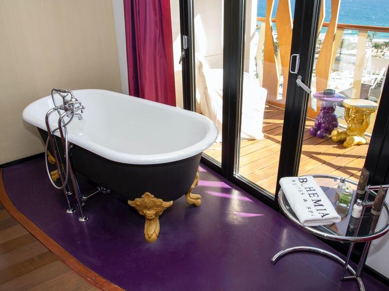 Vasca da bagno autonoma con piedi dorati a forma di leone su pavimento viola vicino a porte in vetro con vista mare.