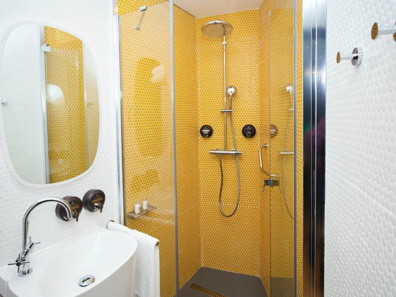 Piccola doccia d'hotel con piastrelle gialle, specchio, lavabo e soffione doccia.