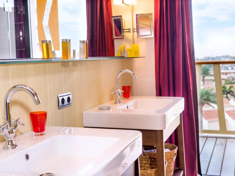 Bagno luminoso con lavandini bianchi, bicchieri rossi e vista su un balcone.