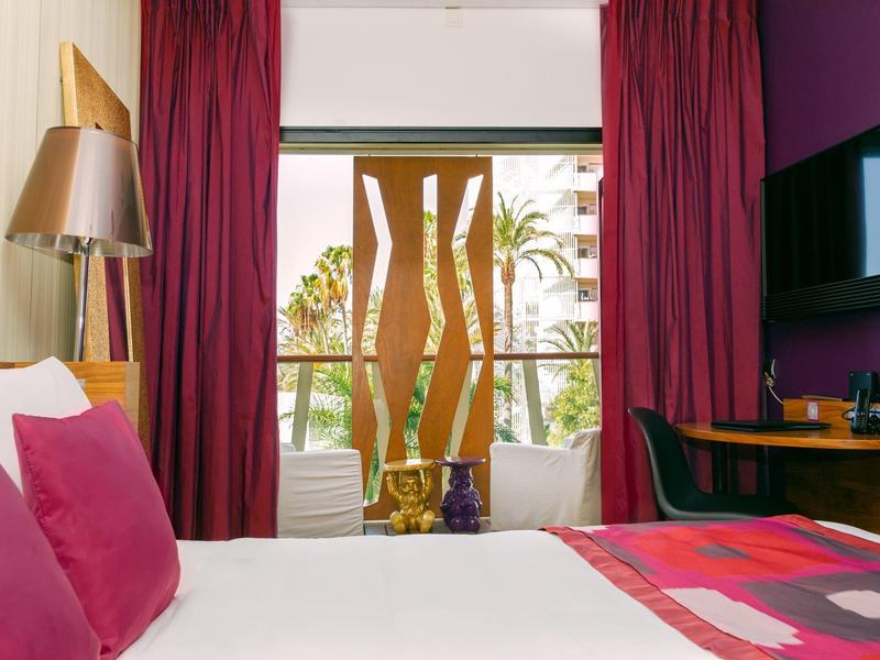 Camera d'hotel con letto grande, tende viola e balcone con vista su palme.