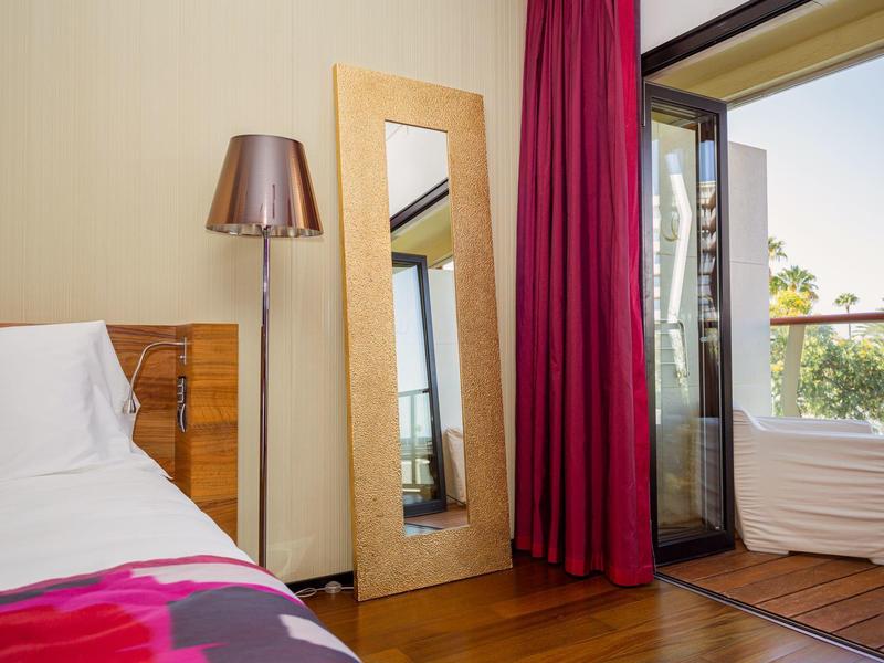 Camera d'hotel luminosa con pavimento in legno, letto, lampada da terra, specchio a figura intera e balcone con ringhiera in vetro.