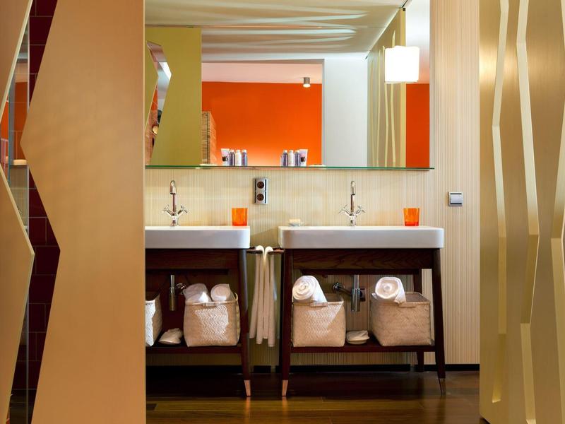 Area bagno moderna in hotel con lavandini, parete arancione e cesti per il bucato sotto il bancone.