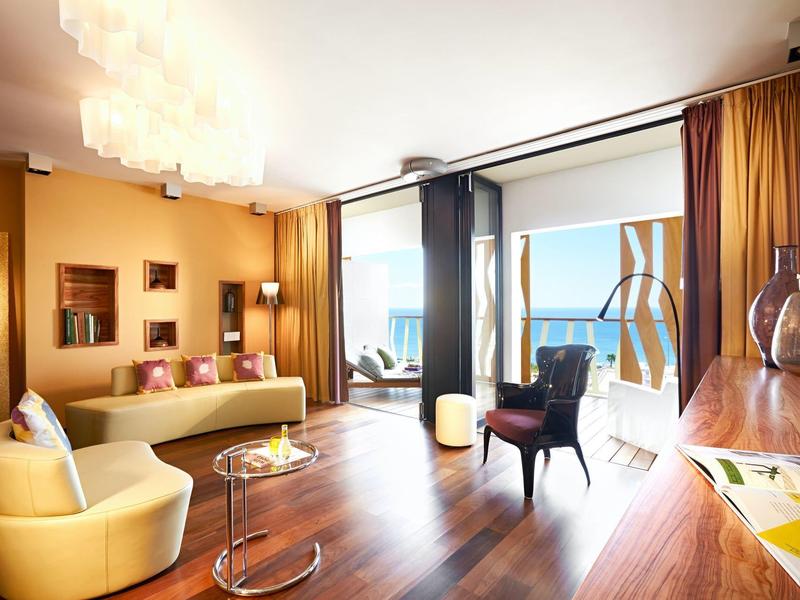 Camera d'hotel luminosa con pavimento in legno, divano giallo, poltrone e vista terrazza sul mare.