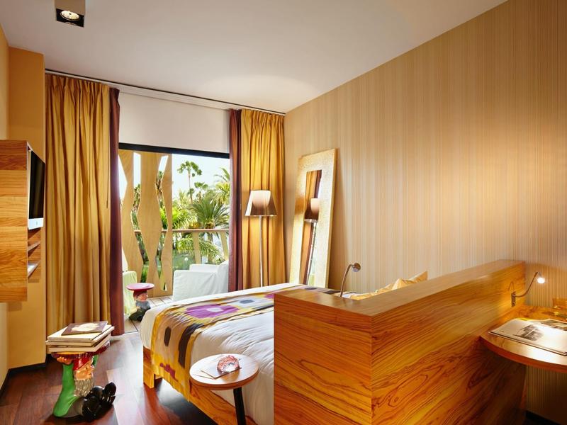 Camera d'hotel luminosa con mobili in legno, letto, sedia e vista sul giardino attraverso grandi finestre.