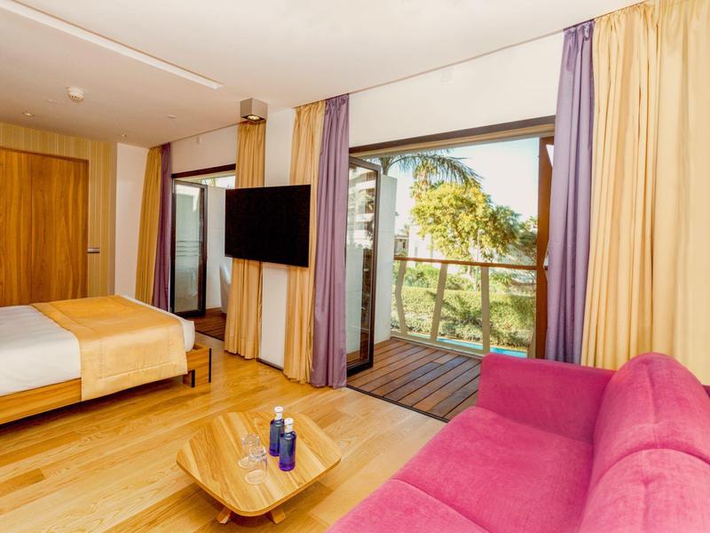 Camera d'hotel moderna con balcone, letto grande, TV a schermo piatto e comodo divano rosa.