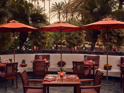 Restaurante al aire libre con sombrillas rojas, mesas y sillas de madera en entorno tropical.