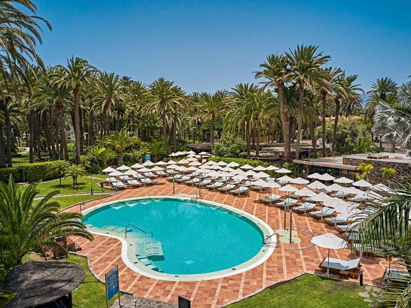 Piscina redonda con muchas tumbonas blancas y palmeras bajo un cielo despejado.