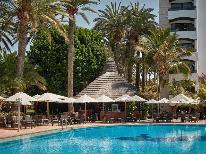 Una piscina con agua azul frente a palmeras y sombrillas en un hotel resort.