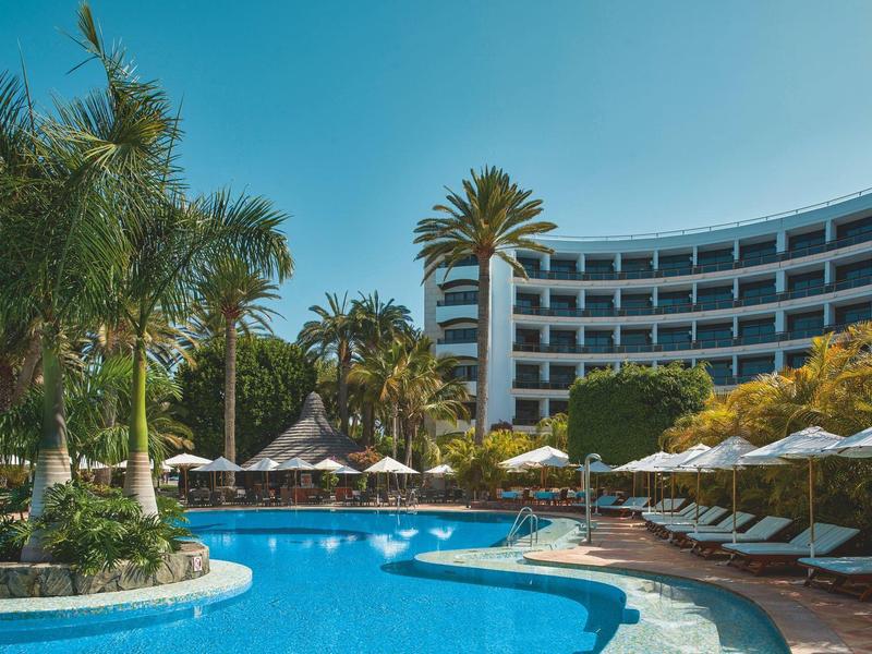 Piscina rodeada de palmeras y tumbonas frente a un edificio de hotel curvado en día soleado