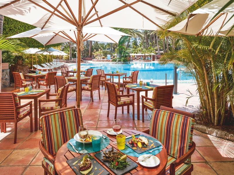 Área de restaurante al aire libre con sombrillas, sillas y mesas junto a una piscina en un entorno tropical.