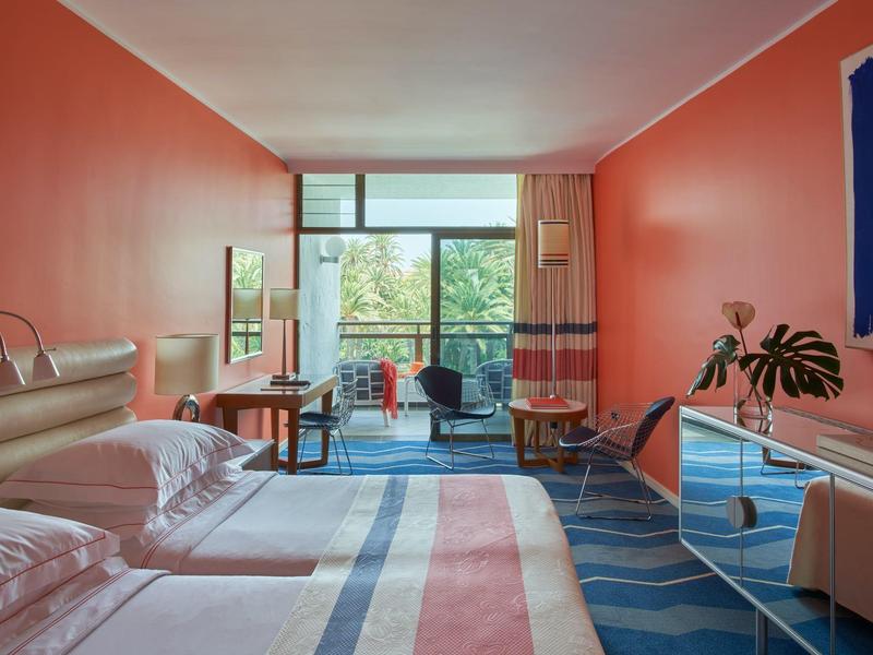 Habitación de hotel moderna con paredes naranjas, cama grande y balcón con vista a la vegetación.