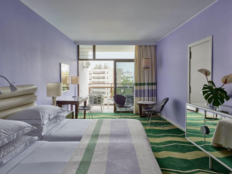 Habitación de hotel moderna con cama doble, alfombra colorida y balcón con vista a la ciudad.