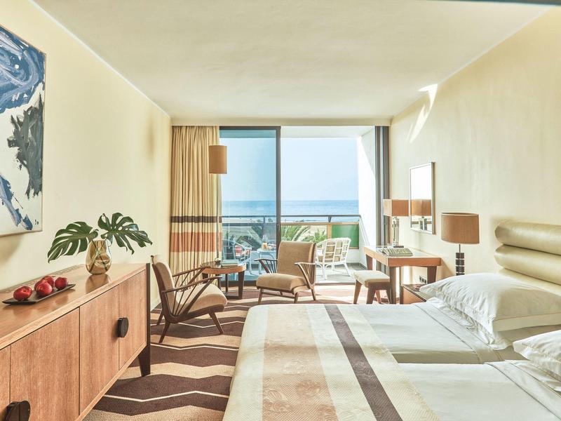 Habitación de hotel luminosa con vista al mar, mobiliario moderno y gran ventanal hacia la terraza.