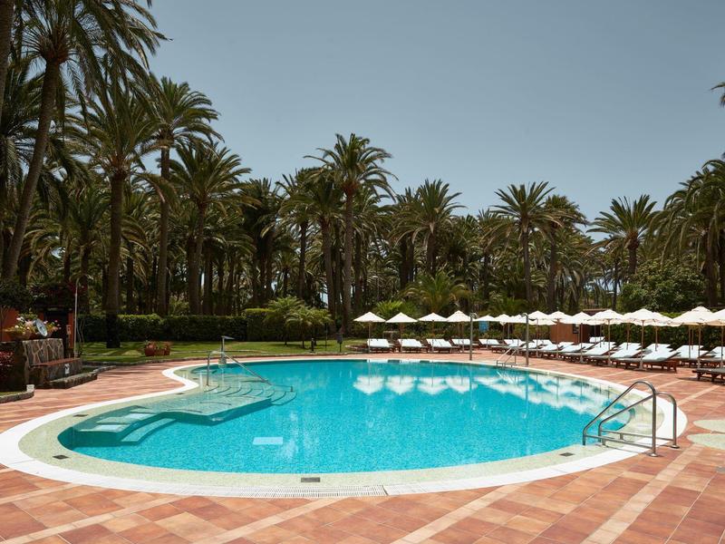 Piscina redonda de hotel con tumbonas bajo sombrillas, rodeada de palmeras y cielo azul.