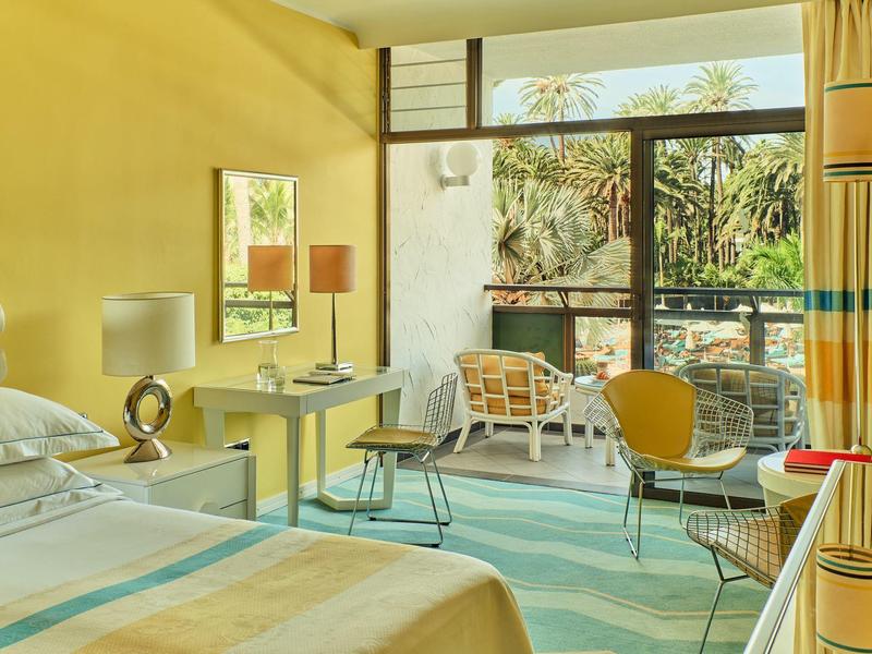 Habitación de hotel luminosa con cama, escritorio, sillas y balcón con vista al jardín.