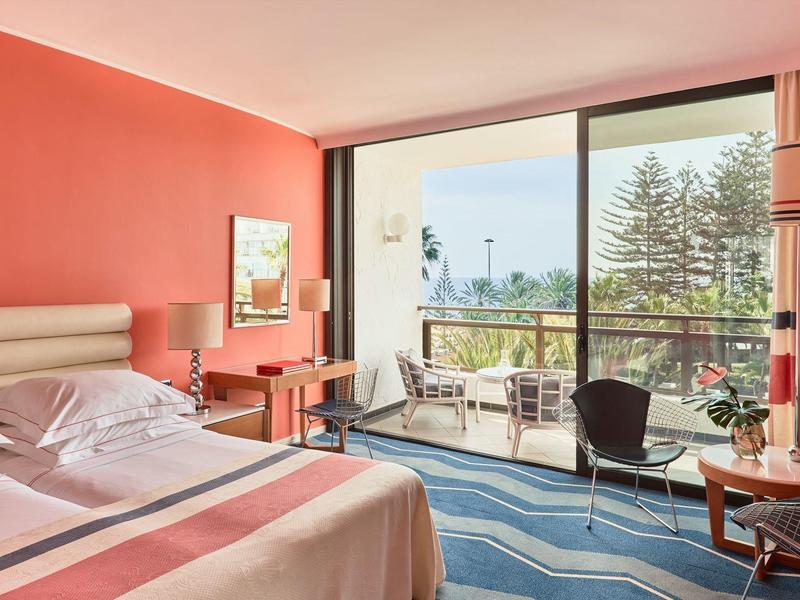 Habitación de hotel luminosa con cama doble, paredes rojas, escritorio y balcón con vista a árboles.