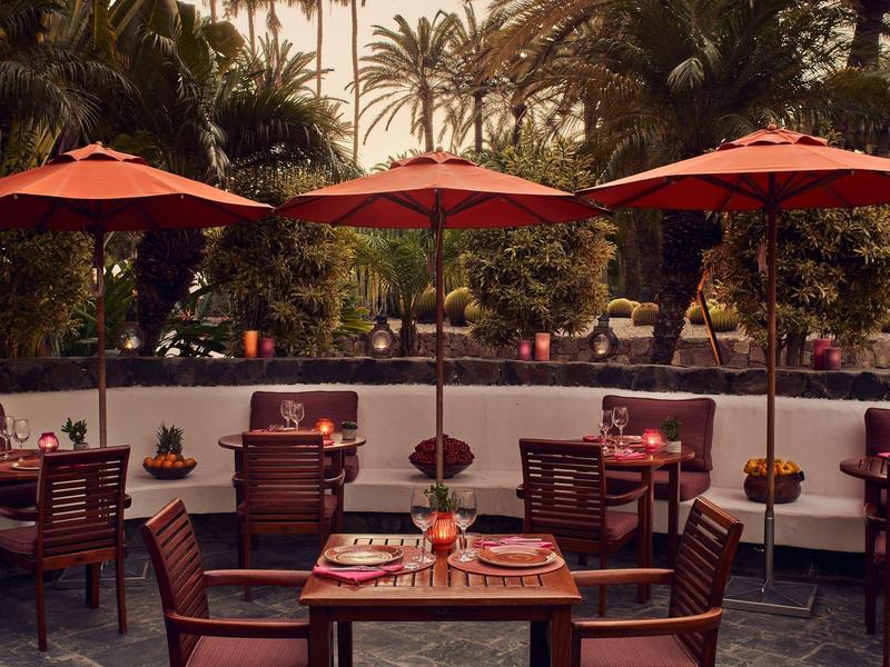 Restaurante al aire libre con sombrillas rojas, mesas y sillas de madera en entorno tropical.