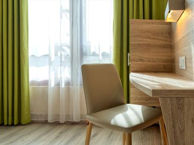 Helles Hotelzimmer mit grünem Vorhang, Schreibtisch und beigem Stuhl.