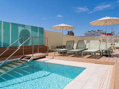 Moderner Hotelpool mit Liegestühlen und Sonnenschirmen auf Holzterrasse bei klarem Himmel.