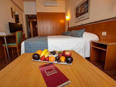 Pohodlný hotelový pokoj s manželskou postelí, dřevěným nábytkem, tácem s ovocem a červeným sešitem na stole.