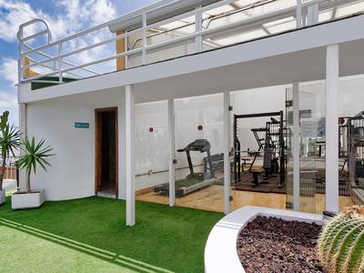 Venkovní fitness prostor se skleněnými stěnami a zeleným umělým trávníkem pod bílou budovou.