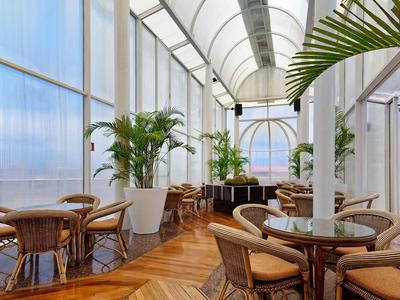 Světla vnitřní terasa s dřevěnými stoly, židlemi, rostlinami a velkými okny s výhledem na město.