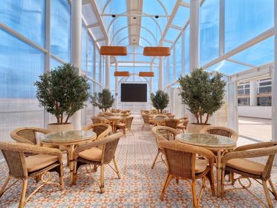 Helle, gläserne Hotel-Lounge mit Naturholz-Möbeln und großen Zimmerpflanzen auf gemustertem Boden.
