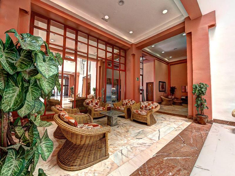 Hotelová lobby s ratanovými křesly, mramorovou podlahou, velkými okny a pokojovými rostlinami.