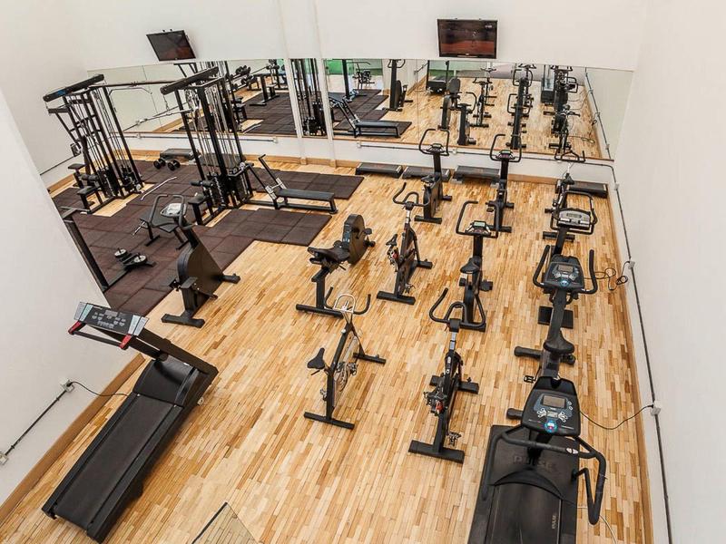 Moderní posilovna s dřevěnou podlahou, kardio stroji a závažími u zrcadlových stěn.
