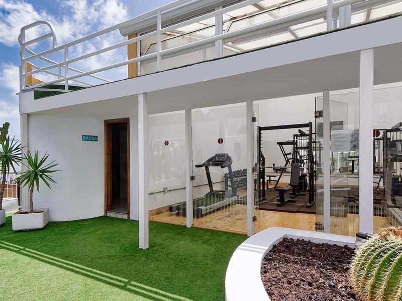 Venkovní fitness prostor se skleněnými stěnami a zeleným umělým trávníkem pod bílou budovou.