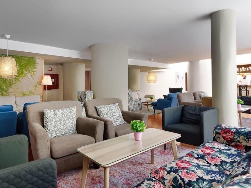 Jasná hotelová hala s květinovými pohovkami, křesly, dřevěnými stoly a dekorativními rostlinami.