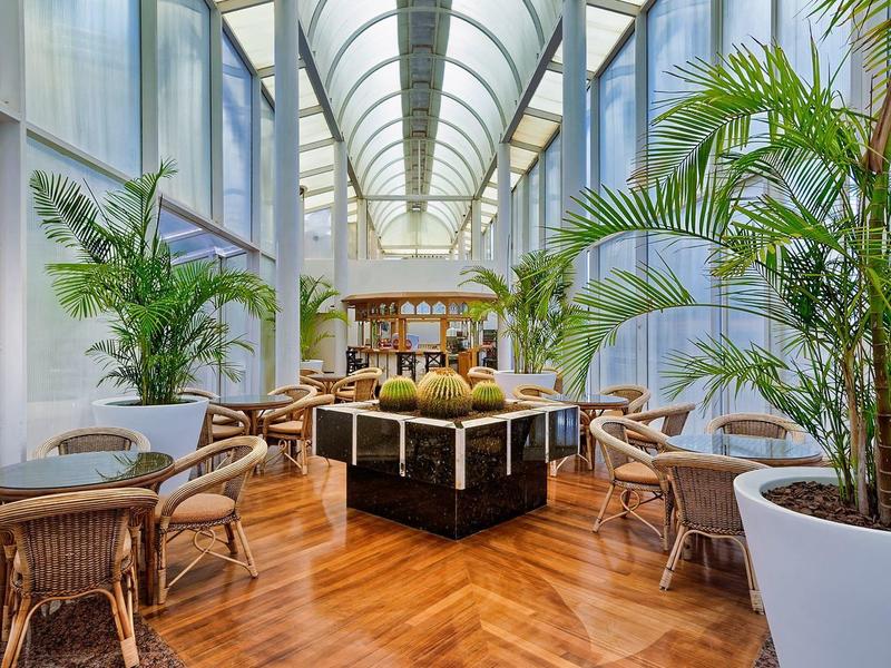 Světlý vnitřní prostor se židlemi z ratanu, dřevěnou podlahou a velkými zelenými rostlinami.