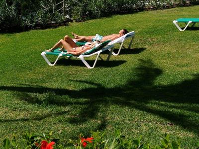 Zwei Personen liegen auf Liegestühlen auf grünem Gras mit roter Blume und Blick aufs Meer.