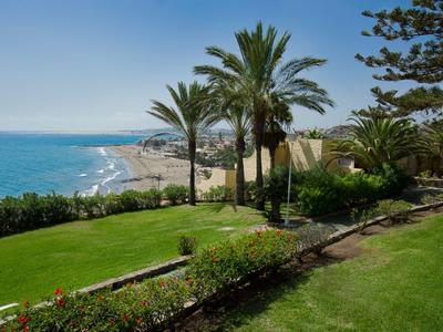 Grüne Gartenanlage mit Palmen und Blick auf einen sonnigen Sandstrand und das blaue Meer.