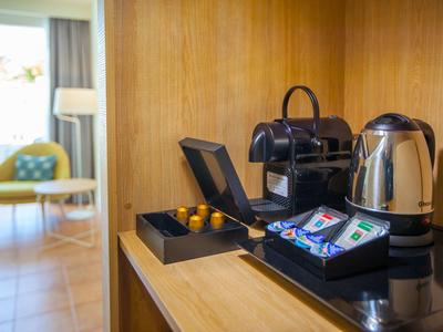 Kaffee- und Teestation mit Kapselmaschine, Wasserkocher und Tassen auf Holztheke in modernem Raum.