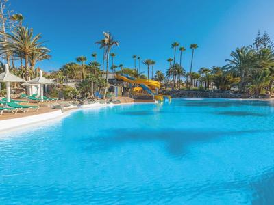 Large blue hotel pool with sun loungers and palm trees under clear sky.