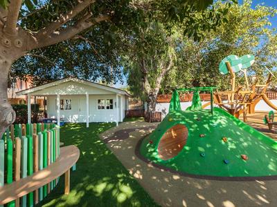 Kinderspielplatz mit Kletterhügel, Schaukel, Holzzäunen und Bäumen unter blauem Himmel.