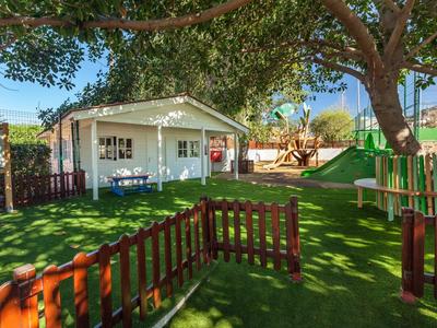 Kinderspielplatz mit grünem Kunstrasen, Holzzaun, Rutsche und weißem Gartenhaus unter Bäumen.